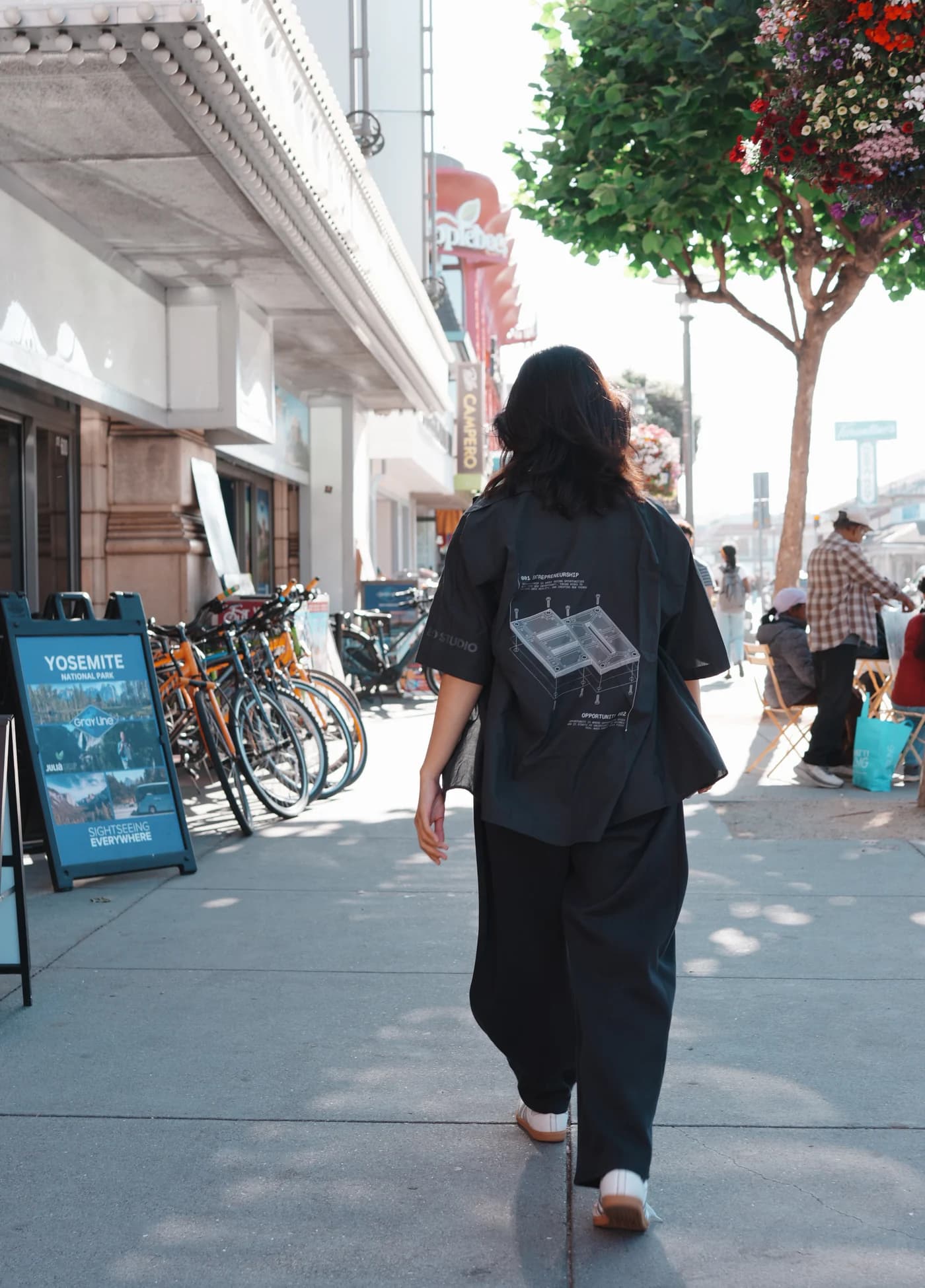 The 2026 EO uniform shirt on a San Francisco street, seen from behind: an exploded-view line drawing across the back, EO STUDIO printed on the sleeve